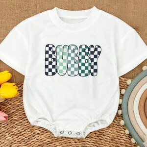 May include: White baby onesie with the word "BUBBY" in a checkered pattern of black, green, and blue. The onesie has short sleeves and snaps at the bottom. The background is a woven texture.