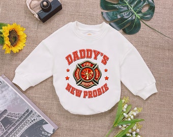 May include: White long-sleeved sweatshirt with a red and black firefighter emblem and the text "Daddy's New Probie".