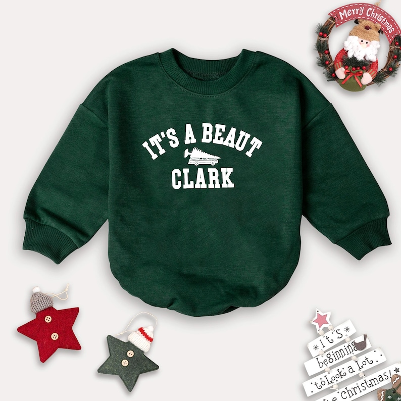 May include: A dark green sweatshirt with the text "IT'S A BEAUT CLARK" in white. The sweatshirt has long sleeves and a round neckline. Christmas decorations, including a wreath and star ornaments, surround the sweatshirt.