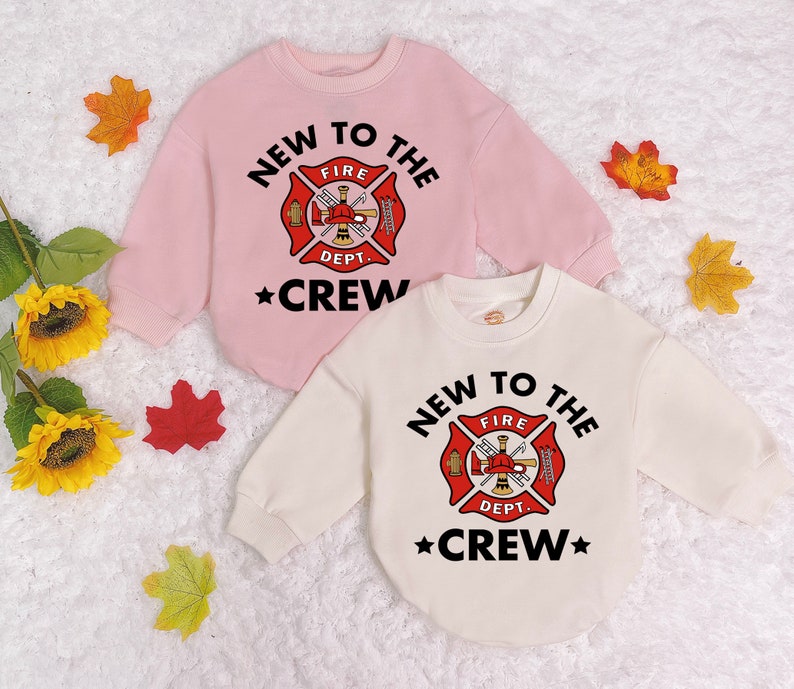 May include: Two long-sleeved shirts with a firefighter theme. The pink shirt says "NEW TO THE FIRE DEPT. *CREW*" and the white shirt says "NEW TO THE FIRE DEPT. *CREW*" with a red firefighter emblem.
