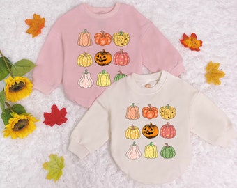 May include: Two pink and cream sweatshirts with a pumpkin and jack-o-lantern design. The sweatshirts have a crew neck and long sleeves. The pumpkins are orange, green, and brown.