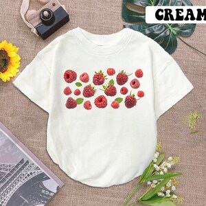 May include: A white short-sleeved t-shirt with a print of red raspberries on the front. The shirt is labeled "CREAM" in the top right corner.