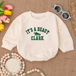 May include: Cream-colored baby onesie with long sleeves. The text "IT'S A BEAUT CLARK" is printed in green across the front, with a Christmas tree graphic. The onesie is on a tan surface, with sunglasses and a beaded necklace.