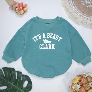 May include: Teal-colored baby sweatshirt with the text "IT'S A BEAUT CLARK" in white block letters. The design includes a graphic of a shoe. The sweatshirt has a crew neck and long sleeves.