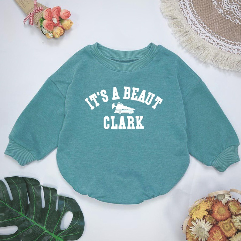 May include: Teal-colored baby sweatshirt with the text "IT'S A BEAUT CLARK" in white block letters. The design includes a graphic of a shoe. The sweatshirt has a crew neck and long sleeves.