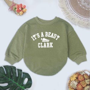 May include: Olive green sweatshirt with the text "IT'S A BEAUT CLARK" in white block letters. The sweatshirt has a crew neck and long sleeves. The graphic includes a stylized image of a vehicle.
