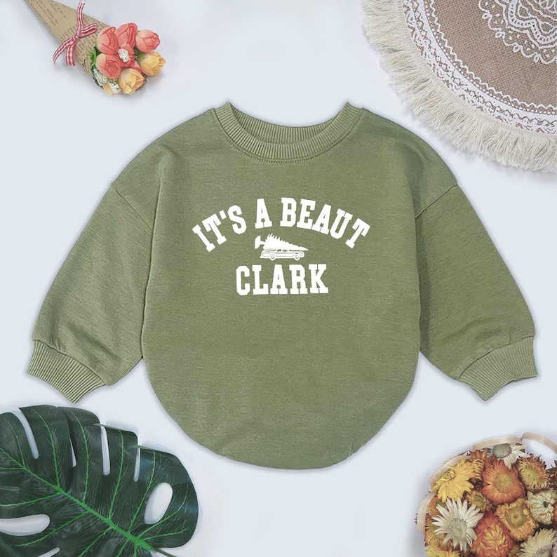 May include: Olive green sweatshirt with the text "IT'S A BEAUT CLARK" in white block letters. The sweatshirt has a crew neck and long sleeves. The graphic includes a stylized image of a vehicle.