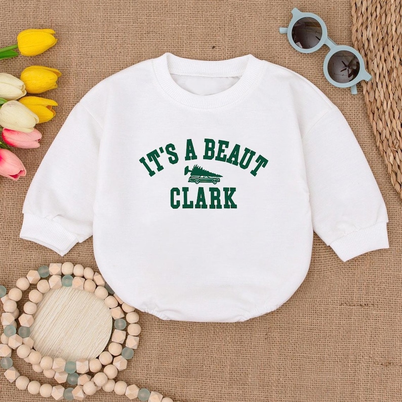 May include: White baby bodysuit with long sleeves. The front features the text "IT'S A BEAUT CLARK" in green, with a graphic of a Christmas tree on a car. The bodysuit is made of a soft material.