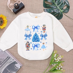 May include: White long-sleeve sweatshirt with a blue and white Christmas design. The design features a Christmas tree, a nutcracker, a ballerina, and blue bows.