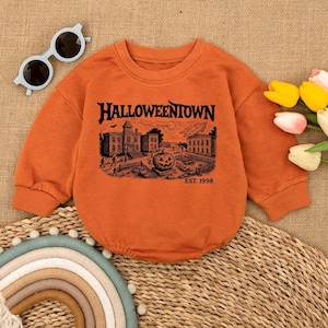 May include: Orange long-sleeved baby romper with the text "Halloweentown" and a black graphic of a spooky town scene with a jack-o'-lantern. The romper also has the text "EST. 1998". Includes a pair of light blue sunglasses.