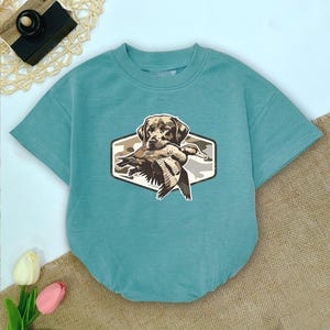 May include: A teal short-sleeved bodysuit with a crew neck. The front features a graphic of a brown Labrador Retriever holding a duck in its mouth, set within a camouflage-patterned hexagon.