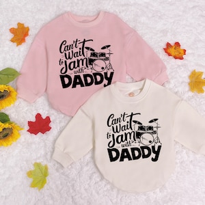 May include: Two pink and white long-sleeved shirts with a black graphic of a drum set and the text "Can't Wait to Jam with Daddy".