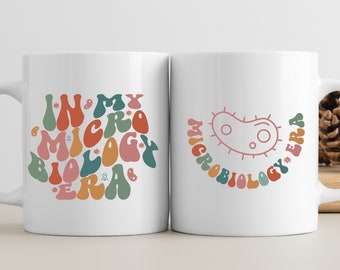 Puede incluir: Dos tazas blancas con un tema de microbiología. Una taza presenta el texto "In My Microbiology Era" en letras coloridas de estilo retro. La otra taza presenta una bacteria rosa estilizada con el texto "Microbiology Era" en letras coloridas de estilo retro.