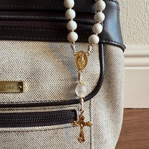 May include: A rosary with white beads, gold-coloured accents, and a gold cross pendant hangs in front of a handbag. The rosary features a gold medallion with a religious figure. The handbag has a neutral fabric with brown leather trim.