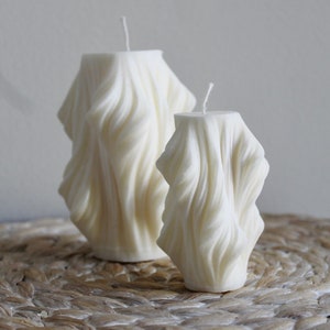 May include: Two ivory-coloured candles with a textured, draped design. The candles are different sizes, with a wick visible on top. They are placed on a woven, circular mat against a neutral background.