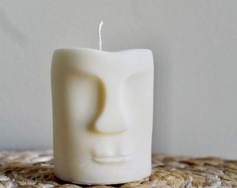 May include: A cream-colored candle shaped like a face with a prominent nose, closed mouth, and subtle eye indentations. The candle has a wick at the top and sits on a woven surface. The candle is a decorative item.