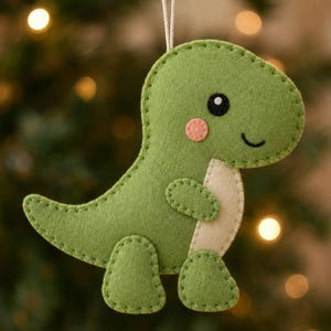 May include: A green felt dinosaur ornament featuring a cream-colored belly, a small pink circular cheek detail, and black stitched eyes and mouth. The dinosaur is constructed with visible blanket stitching along the edges and includes a white string loop.
