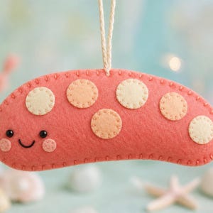 May include: A coral-colored felt ornament shaped like a sea slug with a smiling face, black bead eyes, and pink cheeks. The body features several cream and light orange circular felt spots stitched onto the surface. A white string is attached to the top.