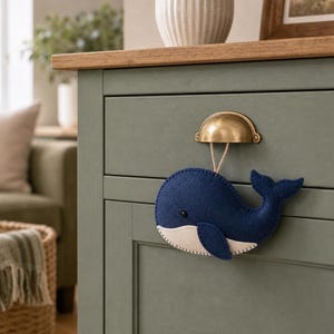 May include: A dark blue felt whale ornament with a light cream-colored underbelly and a small black eye. The whale is suspended by a thin tan string from a brass-colored metal drawer pull attached to a sage green cabinet drawer front.