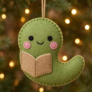 May include: A green felt ghost ornament featuring a tan open book held in the center. The ghost has two black circular eyes, a small stitched smile, and two pink circular cheeks. It hangs from a brown twine loop at the top of the head.
