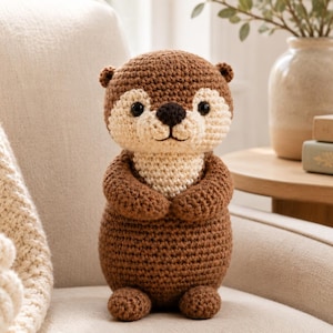 May include: A handmade crochet otter plush toy. The otter is brown with a cream-colored face and chest, black eyes, and a black nose. It is sitting on a cream-colored chair. The otter has small arms and feet. The background includes a vase with greenery and a stack of books.