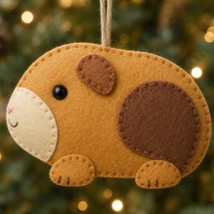 May include: A felt guinea pig ornament in shades of tan, cream, and dark brown. The animal figure features a black bead eye, a stitched pink nose, and a small smile. It is suspended by a thin jute twine loop and has visible blanket stitching.