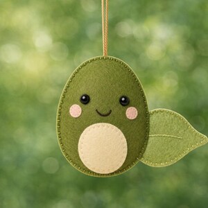 May include: A felt avocado ornament featuring a dark green oval body, a light cream-colored belly patch, and a single green leaf attached to the side. The face includes two black bead eyes, a stitched smile, and two small pink felt circles for cheeks.