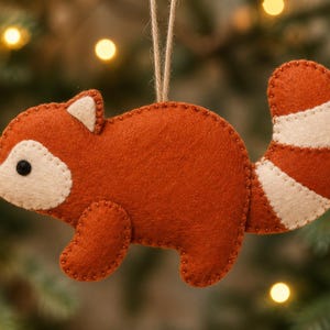 May include: A felt red panda ornament featuring a rust-orange body with cream-colored accents on the face, ears, and striped tail. The animal figure has a small black nose and eye, with visible stitching along the edges and a jute twine loop for hanging.