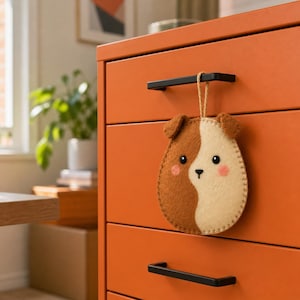 May include: A felt ornament shaped like a dog head with a split brown and cream colour pattern, featuring small ears, black eyes, a black nose, and pink cheeks. The ornament hangs from a thin jute string attached to a black drawer handle.