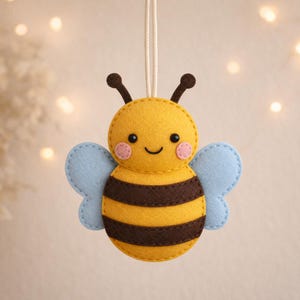 May include: A felt bee ornament featuring a yellow and brown striped body, light blue wings, and two antennae. The bee has a stitched face with black eyes, a small smile, and pink circular cheeks. A white string is attached to the top for hanging.