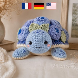 May include: A handmade crochet turtle in shades of blue, with a light blue head and shell adorned with darker blue accents. The turtle has a smiling face, pink cheeks, and green leaf details. The toy is on a white surface.