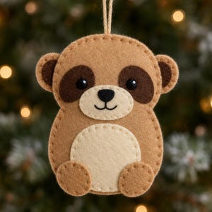 May include: A felt meerkat ornament featuring a light brown body, dark brown eye patches, a cream-colored belly, and black stitched facial details. The ornament is suspended by a thin twine loop and has visible blanket stitching along the edges.