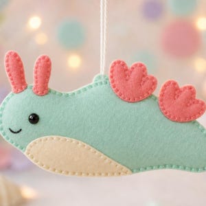 May include: A felt sea slug ornament in mint green with a cream-coloured belly, two coral-pink rhinophores, and two coral-pink dorsal gills. The creature features a stitched outline, a small black bead eye, and a simple embroidered smile.