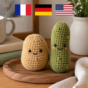 May include: A crocheted potato and pickle with smiling faces sit on a wooden tray. The potato is tan, and the pickle is green. A white mug, books, and a vase with greenery are in the background.