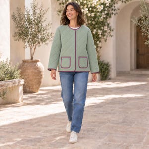 Può includere: Una giacca trapuntata verde salvia con scollo rotondo, maniche lunghe e due tasche applicate sul davanti. La giacca è rifinita con un bordo a contrasto viola scuro lungo i profili e le tasche, abbinata a jeans blu e scarpe da ginnastica bianche.