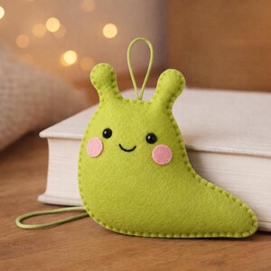 May include: A lime green felt slug ornament featuring two small black eyes, a stitched black smile, and two round pink felt cheeks. The slug has a loop at the top for hanging and is constructed with visible stitching along the outer edges of the felt.