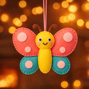 May include: A felt butterfly ornament featuring a yellow body, pink upper wings with light pink circles, and teal lower wings with light pink circles. The butterfly has a smiling face with black eyes and pink cheeks, brown antennae, and a pink hanging string.