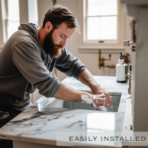 Op de afbeelding: Een man wast zijn handen in een witte marmeren gootsteen met een wit aanrecht. De gootsteen maakt deel uit van een badkamermeubel met een grijze kast. De tekst "EASILY INSTALLED" is zichtbaar onderaan de afbeelding.
