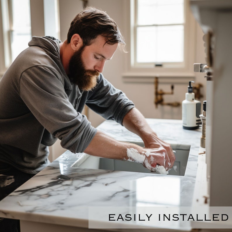 Op de afbeelding: Een man wast zijn handen in een witte marmeren gootsteen met een wit aanrecht. De gootsteen maakt deel uit van een badkamermeubel met een grijze kast. De tekst "EASILY INSTALLED" is zichtbaar onderaan de afbeelding.