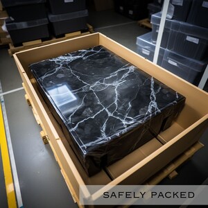 Op de afbeelding: Een zwart marmeren tafelblad is veilig verpakt in een kartonnen doos. De doos staat op een houten pallet. De tekst "SAFELY PACKED" is onderaan de afbeelding gedrukt.