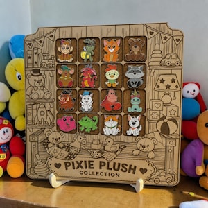May include: A wooden display board featuring sixteen colorful character enamel pins arranged in a grid. The board is engraved with toy-themed illustrations and the text PIXIE PLUSH COLLECTION. It stands on two white plastic feet on a surface.