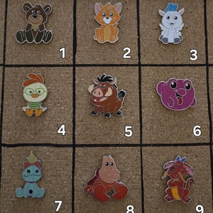 May include: Nine colorful enamel pins featuring cartoon characters, including a bear, cat, goat, chicken, warthog, frog, Stitch, a hippo, and a dragon. Each pin has a black outline and is set against a brown background.