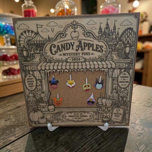 May include: A square wooden display board featuring a laser-engraved candy shop illustration with the text Candy Apples Mystery Pins 2024. Six enamel pins shaped like candy apples are attached to the cork center, including character-themed designs.