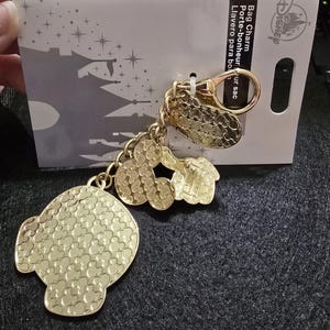Puede incluir: Charm de bolso de Disney dorado con m&uacute;ltiples dijes, incluyendo una cabeza grande y texturizada de Mickey Mouse, formas m&aacute;s peque&ntilde;as de Mickey Mouse y un dije rectangular. El charm est&aacute; unido a un cierre dorado y a una tarjeta blanca con el logo de Disney.