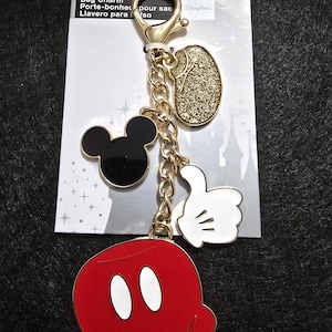 Puede incluir: Un charm de bolso dorado con dijes tem&aacute;ticos de Mickey Mouse. El charm incluye una cabeza de Mickey Mouse, un &oacute;valo dorado con purpurina, una mano enguantada blanca con el pulgar hacia arriba y un charm de pantalones cortos rojos de Mickey Mouse.