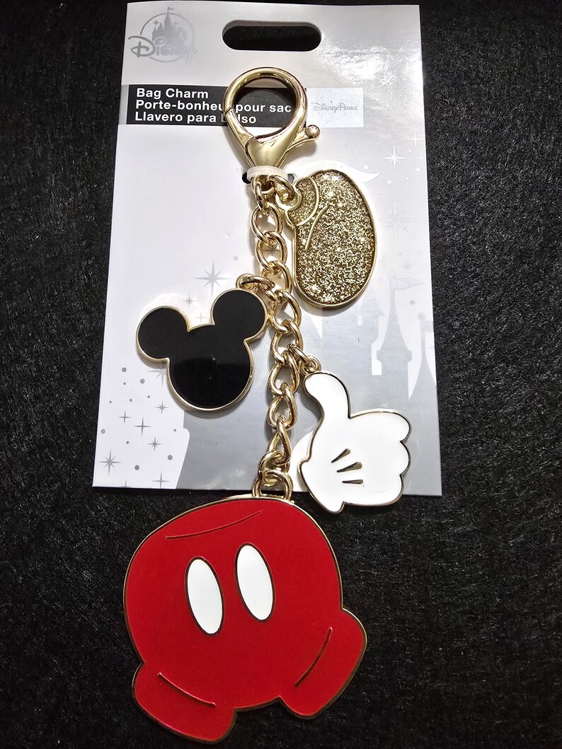 Puede incluir: Un charm de bolso dorado con dijes tem&aacute;ticos de Mickey Mouse. El charm incluye una cabeza de Mickey Mouse, un &oacute;valo dorado con purpurina, una mano enguantada blanca con el pulgar hacia arriba y un charm de pantalones cortos rojos de Mickey Mouse.