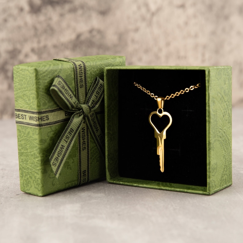 Puede incluir: Un collar de llave de oro con un colgante de coraz&oacute;n con forma de ojo de cerradura, presentado en una caja de regalo verde con una cinta negra y las palabras "Mejores deseos" impresas en la caja.