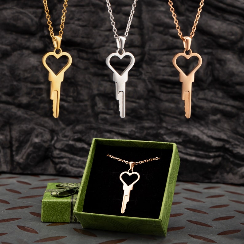 Puede incluir: Tres collares de oro, plata y oro rosa con colgantes de llave en forma de coraz&oacute;n. Los collares se muestran sobre un fondo negro. Un cuarto collar con un colgante de llave en forma de coraz&oacute;n de oro rosa est&aacute; en una caja de regalo verde.