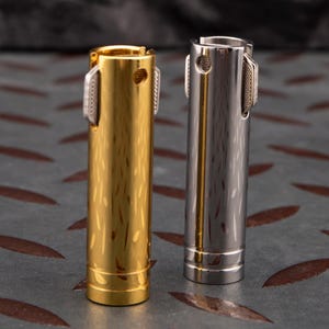 Pode incluir: Dois isqueiros metálicos cilíndricos, um dourado e outro prateado, com acabamento texturizado. Ambos os isqueiros têm um pequeno botão redondo prateado no lado.