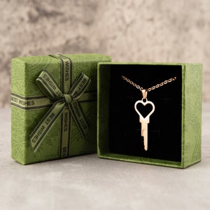 Puede incluir: Un collar de llave de coraz&oacute;n de oro en una caja de regalo verde con interior de terciopelo negro. La caja est&aacute; decorada con una cinta verde y las palabras "Mejores deseos" impresas en ella.